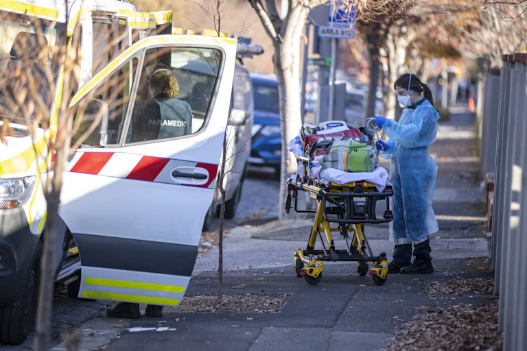 An ambulance worker in PPE stands next to a gurney on a pavement.