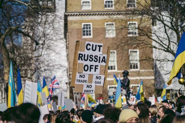 Crowd of protestors with British and Ukraine flags and a sign that says cut Russia off: no trade, no gas, no swift.