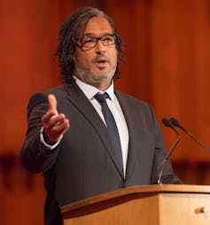 A bi-racial man in a suit at a podium speaking into a mic.
