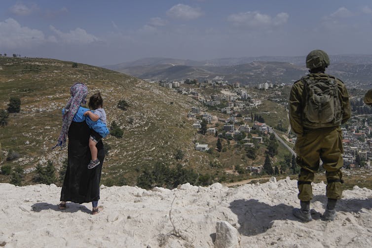 A soldier in battle fatigues stands on a hill next to a woman holding a child.