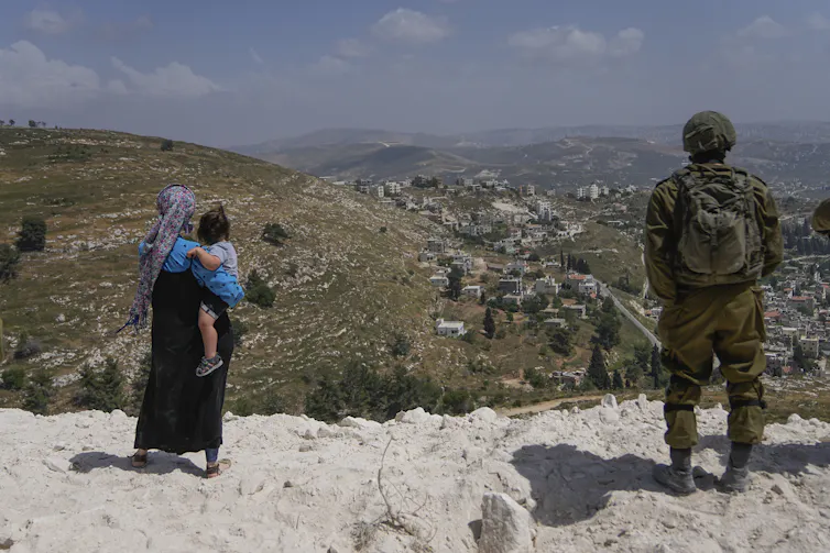 A soldier in battle fatigues stands on a hill next to a woman holding a child.