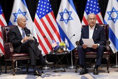 Two men sit and talk with Israeli and U.S. flags behind them.