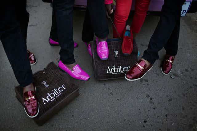 Five sets of legs displaying glossy shoes in red and bright pink, stepping on ashoe boxes with the words "Arbiter - Made in Italy" on them.