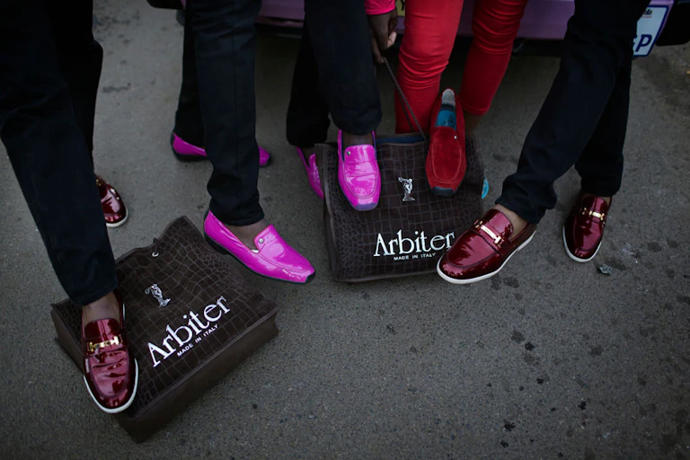 Five sets of legs displaying glossy shoes in red and bright pink, stepping on ashoe boxes with the words "Arbiter - Made in Italy" on them.