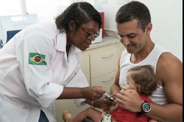 Enfermeira aplica vacina em uma criança pequena, nos braços do pai
