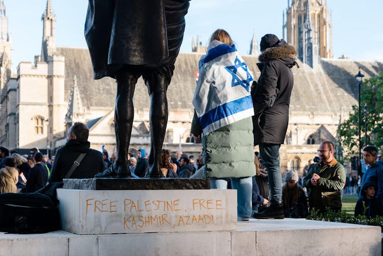 A woman wearing an Israeli flag stands next to a statue in Parliament Square, behind her some graffiti reads 