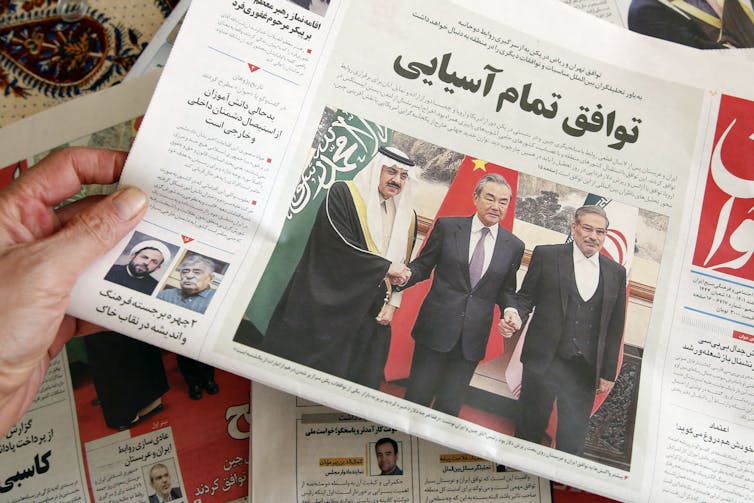 A hand holds a newspaper with Arabic writing and a picture of three men, one wearing traditional Saudi dress.