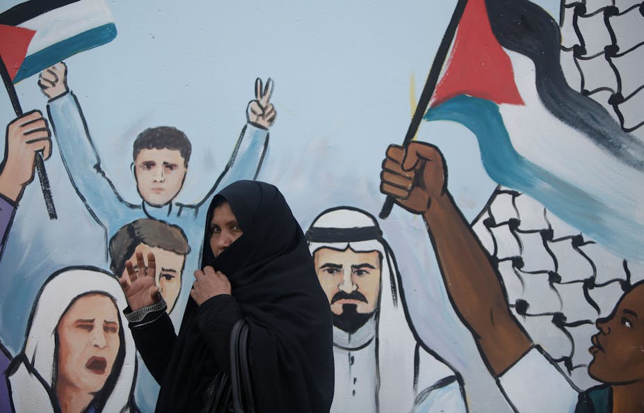 A woman in a headscarf walks past a mural of people flashing 'V' peace signs and holding Palestinian flags.