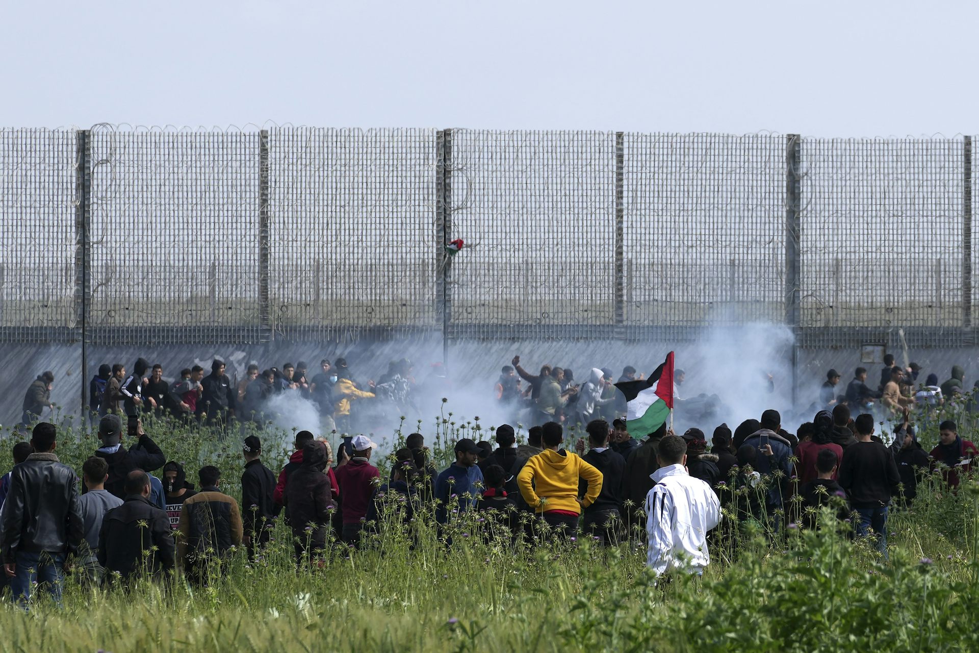 People gather by a large chain-link fence amid clouds of teargas.