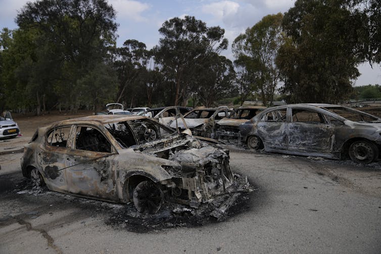 Destroyed, burnt-out cars in a dusty parking lot.
