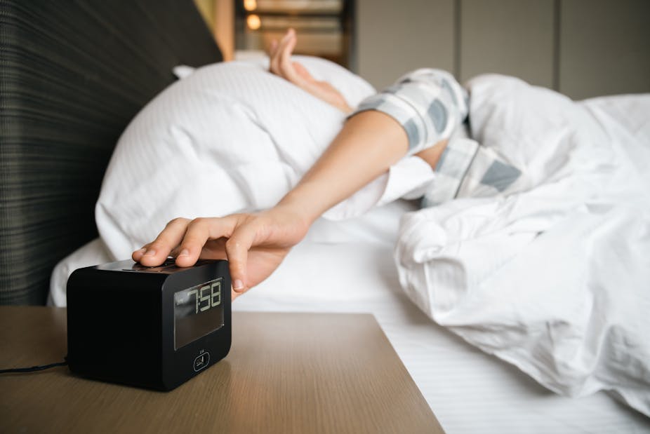 A person lying in bed hits snooze on their alarm clock.