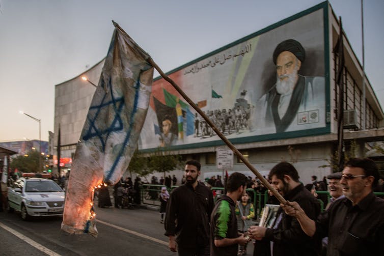 Men stand on a city street with a police car nearby and burn a drawn Israeli flag. Behind them is a large billboard of a man with a white beard and black hat.