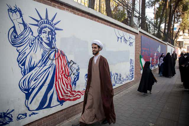 A man wearing a brown robe and white head cover walks along a wall with a large white mural showing the Statue of Liberty with its hand taken off. 