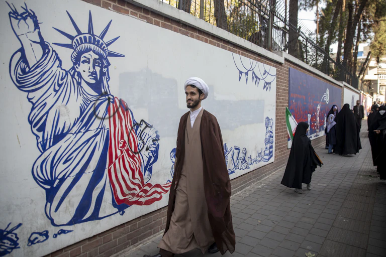 A man wearing a brown robe and white head cover walks along a wall with a large white mural showing the Statue of Liberty with its hand taken off.