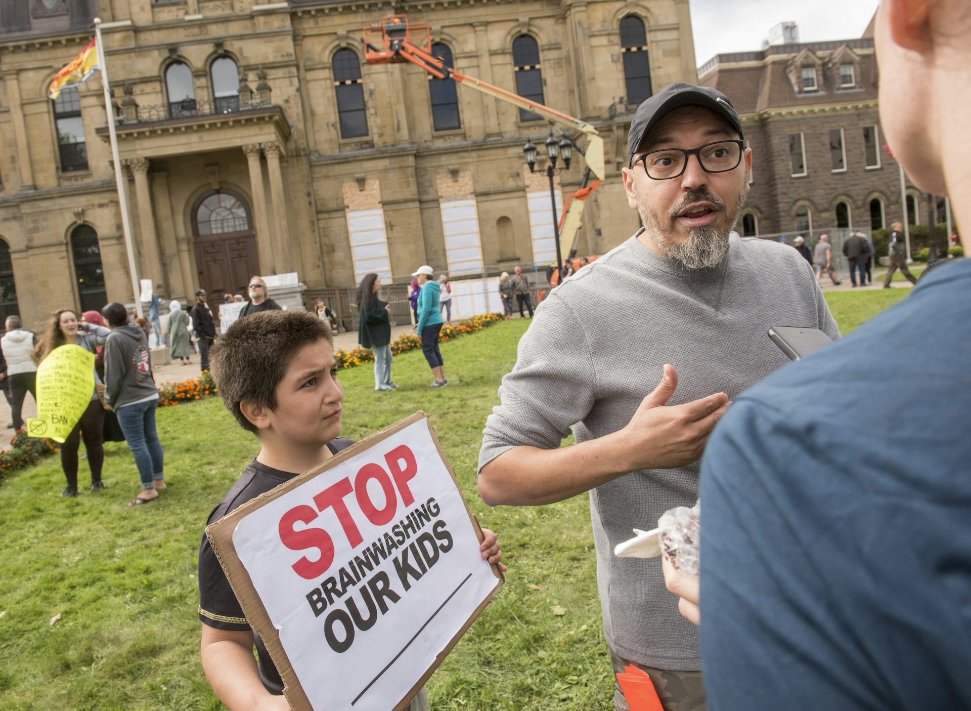 A man speaks to reporters. A young boy beside him carries a sign: stop brainwashing our kids.
