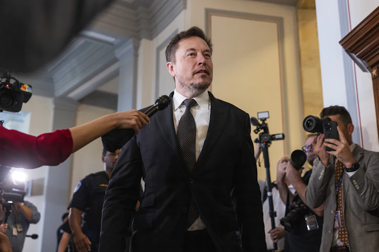 Elon Musk speaking to reporters, people with cameras and microphones.