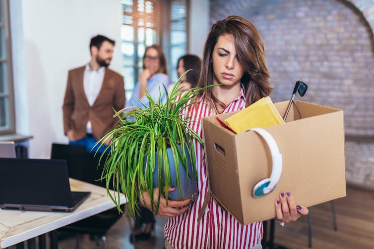 Woman with box of belongings and plant, looking annoyed, people talking in the background.