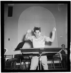 A man in front of a lectern with a baton jumps up and down energetically.