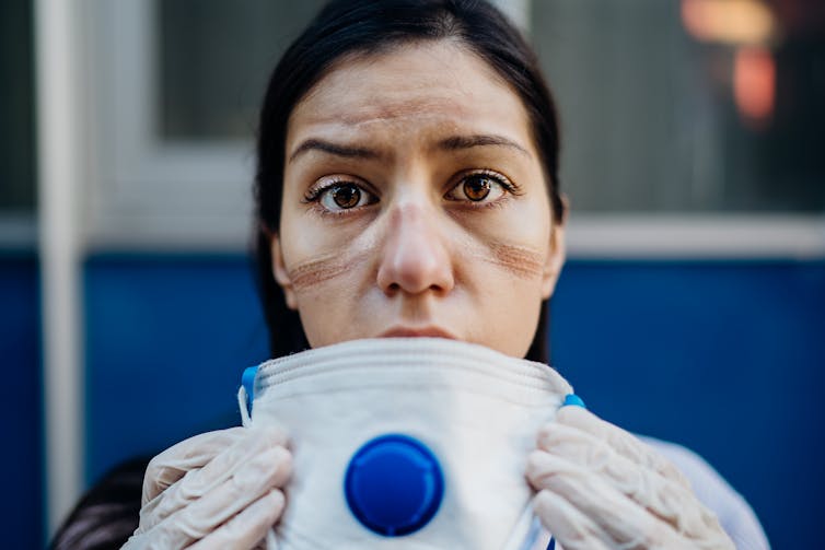 A woman in personal protective equipment removing a face mask looking exhausted