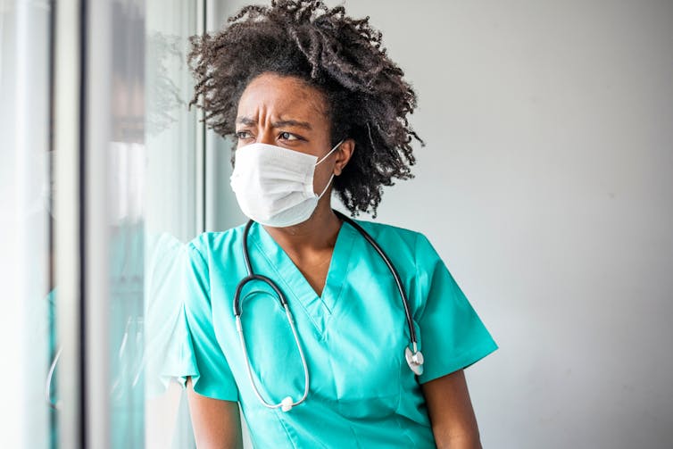 A woman in scrubs and a face mask looking out a window with a furrowed brow