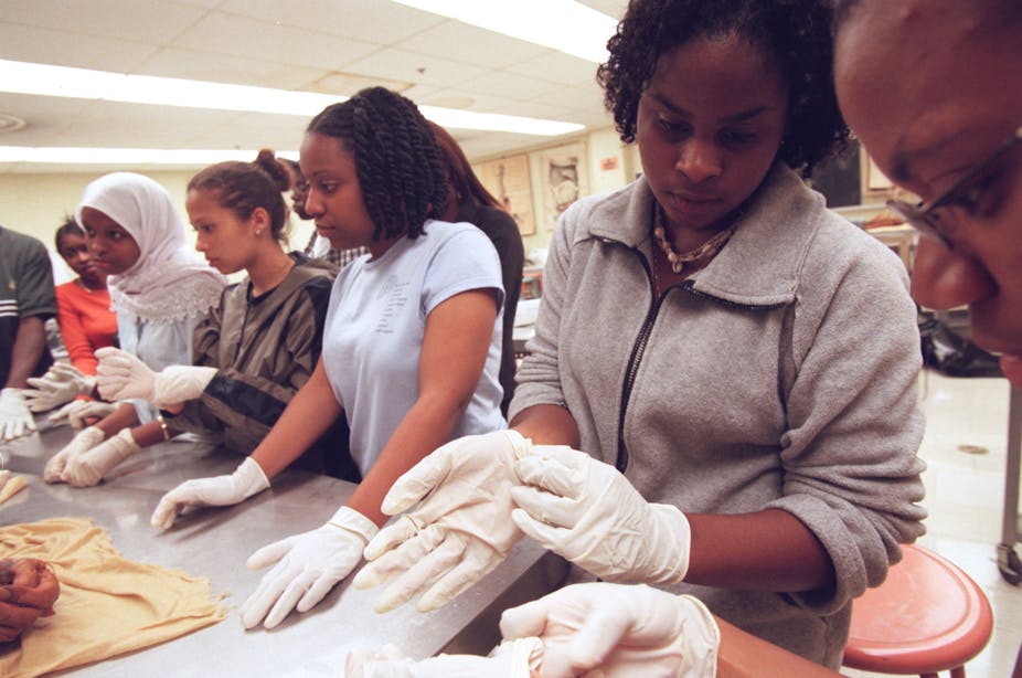 Several young women standing in a row, hands in white gloves, listening intently.