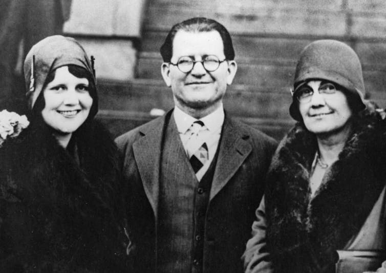 Black and white photo of smiling man with eyeglasses, flanked by two women.