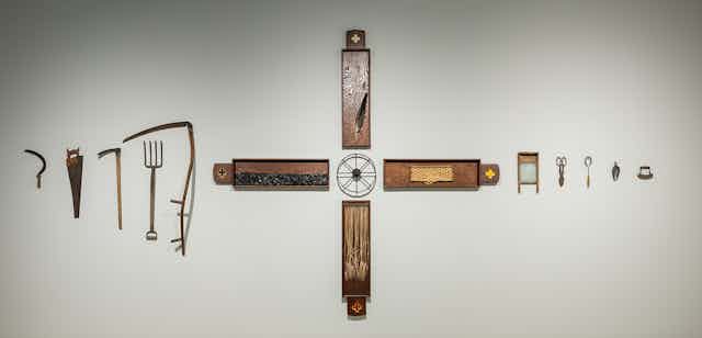 A scultpture installation seen on a gallery wall showing various objects in the shape of a cross with objects related to manual labour alongside the horizontal axis, such as a saw, pitchfork, washing board and scissors.