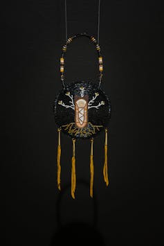 a circular frame holds what appears to show a beading of a child with branches and roots, and fringes of deer hide hang along the bottom of the circular frame, against a black wall, with