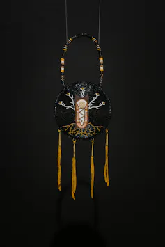 a circular frame holds what appears to show a beading of a child with branches and roots, and fringes of deer hide hang along the bottom of the circular frame, against a black wall, with