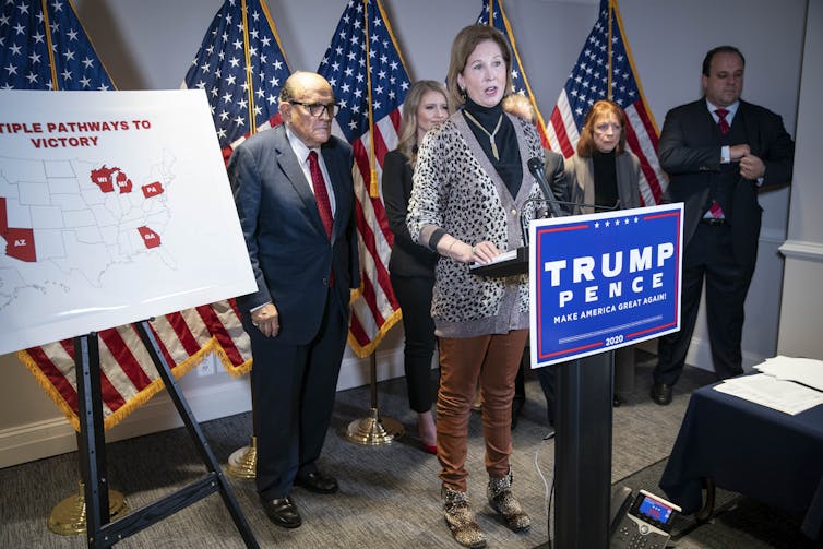 A woman in a suit stands at a lectern labeled 'Trump Pence,' with other people in suits behind her.