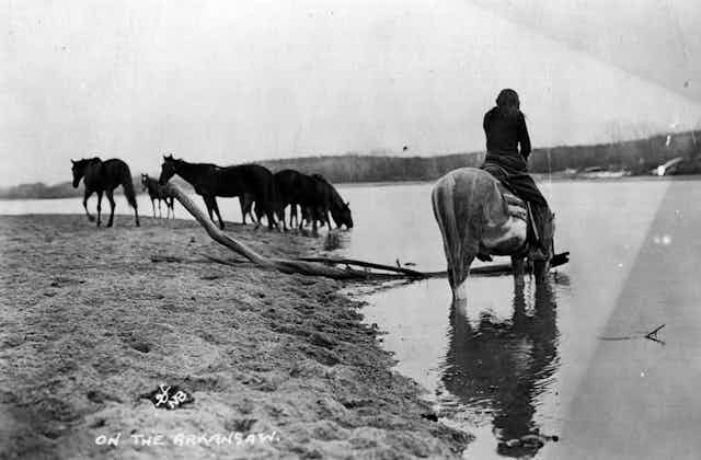 Old photograph of man sitting on a horse that's drinking from a river.