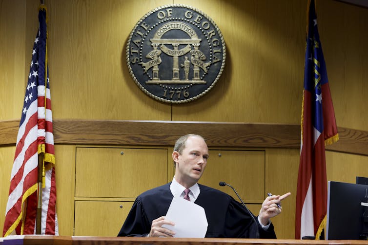 A man in a black robe sitting in front of an American flag and another flag, holding a piece of paper and pointing.