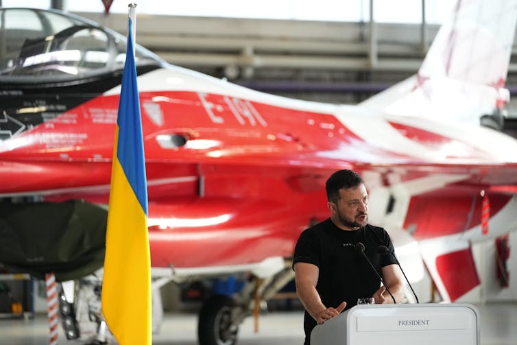 A man in a black t-shirt stands in front of a red-and-white fighter jet. A blue and yellow flag is beside him.