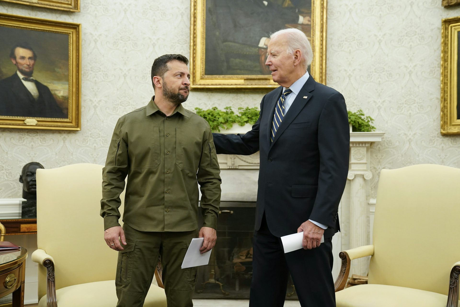 A younger dark-haired man in a green military shirt speaks to an older man in a suit in an ornate office.