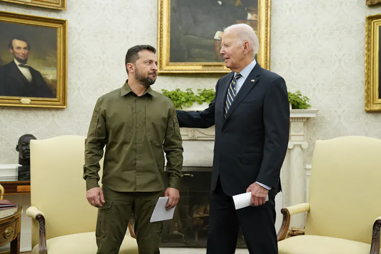 A younger dark-haired man in a green military shirt speaks to an older man in a suit in an ornate office.