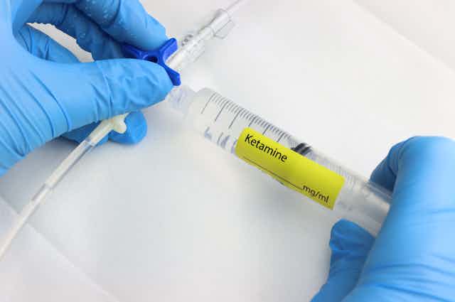 Gloved hands holding a tube of clear liquid labeled as ketamine.