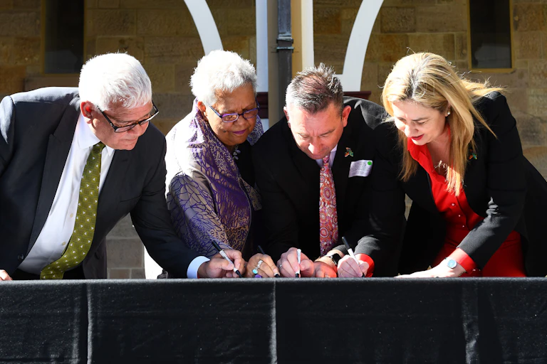 Four people lean over a table signing a document.