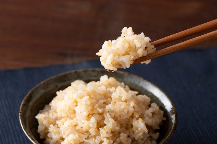 A dark bowl with clumpy tan coloured rice being picked up with wooden chopsticks