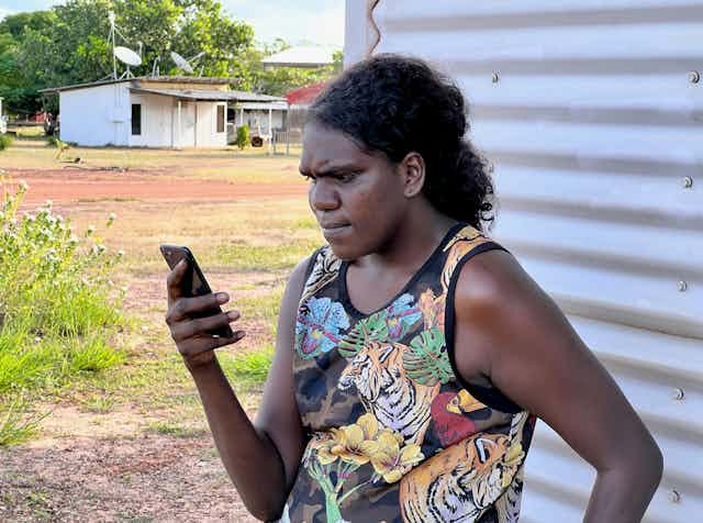 A photo of an Indigenous woman standing outdoors looking at her phone.