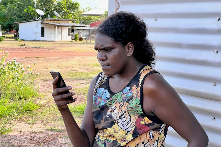 A photo of an Indigenous woman standing outdoors looking at her phone.