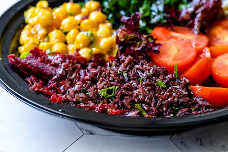 Close-up of a salad on a dark plate with chickpeas, tomato slices and purple rice in the foreground