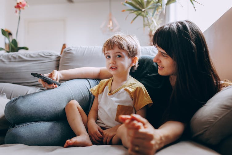 A woman lies on the couch with a remote, and a young child.