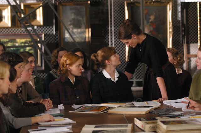 Un grupo de mujeres jóvenes se sientan alrededor de una mesa llena de libros mientras una se encara con otra mujer mayor que ellas que la mira de pie.