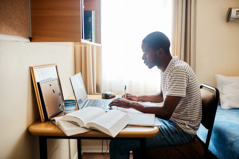 A photograph of a young man sitting at a desk, typing on a laptop. There are books open alongside him and he is holding a pen.