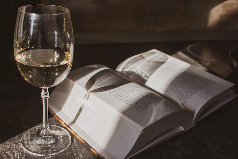 A glass of white wine sits next to a thick open book.