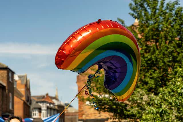 A rainbow shaped and coloured balloon flies in a street. 
