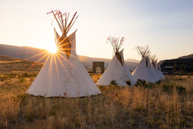 Several teepees stand in a row as the sun rises over a prairie.