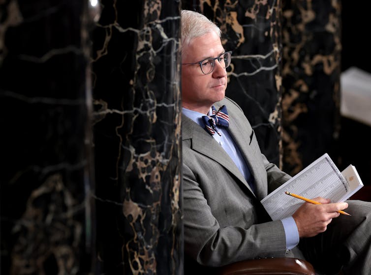 A white haired man in a gray suit and bow tie sits and listens.