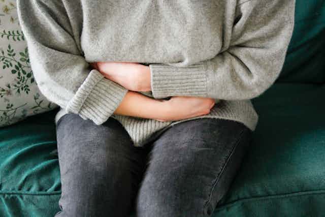 Person in gray sweater and black jeans hugging their torso in pain.