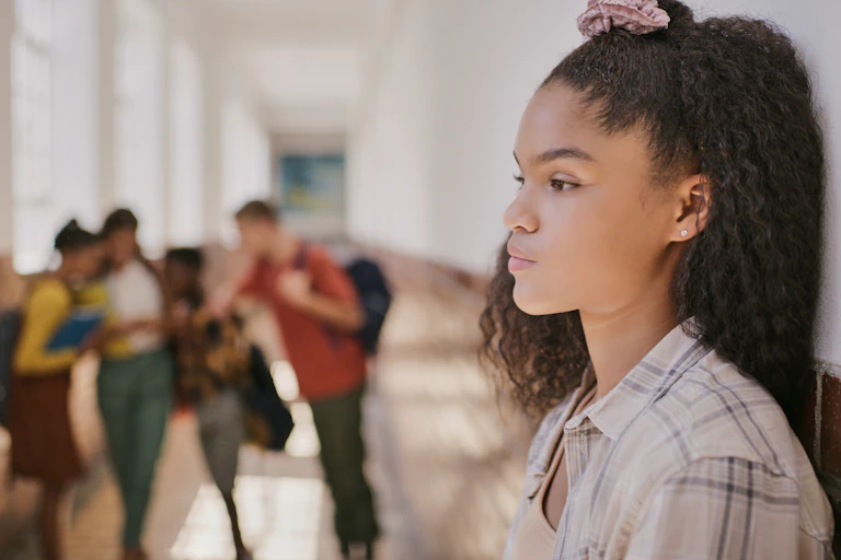 A dejected-looking teenage girl stares off into the distance as her classmates speak disparagingly of her.